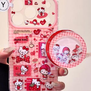 Puede incluir: Funda de teléfono rosa y transparente con diseños de Hello Kitty. La funda tiene un fondo a cuadros rosa con varias ilustraciones de Hello Kitty, corazones y texto. También es visible una funda redonda y transparente separada con figuras de Hello Kitty.