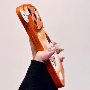 May include: Orange phone case with a cat design. The case has a glossy finish and features a cartoon cat face with black whiskers and a white belly. The case is designed to fit a smartphone and has cutouts for the camera and buttons.
