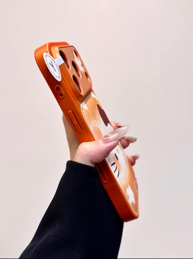 May include: Orange phone case with a cat design. The case has a glossy finish and features a cartoon cat face with black whiskers and a white belly. The case is designed to fit a smartphone and has cutouts for the camera and buttons.
