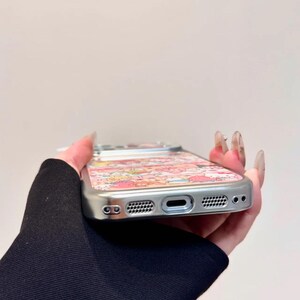 K&ouml;nnte beinhalten: Silberne Handyh&uuml;lle mit einem farbenfrohen Muster aus Zeichentrickfiguren. Die H&uuml;lle hat einen Schutzrahmen und Aussparungen f&uuml;r Kamera, Lautsprecher und Ladeanschluss. Das Design weist rosa, rote und gelbe Farbt&ouml;ne auf.