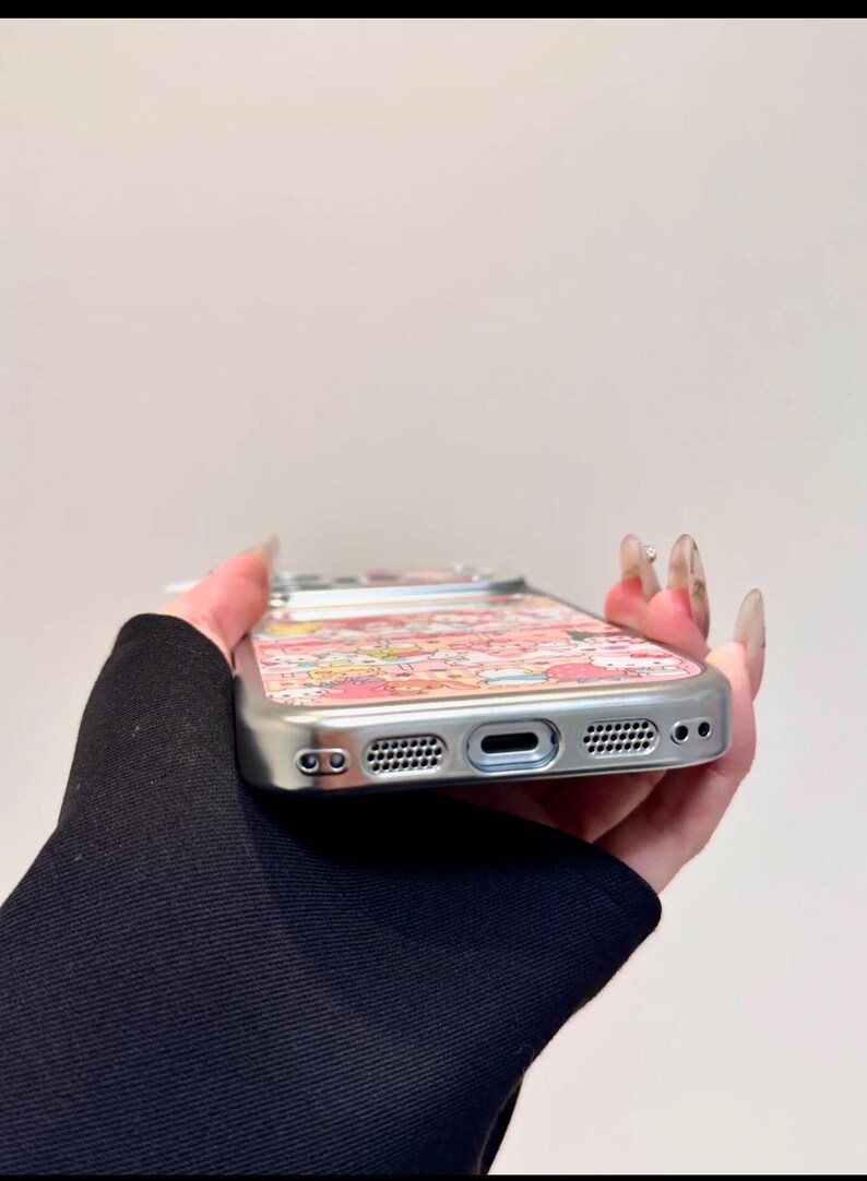 K&ouml;nnte beinhalten: Silberne Handyh&uuml;lle mit einem farbenfrohen Muster aus Zeichentrickfiguren. Die H&uuml;lle hat einen Schutzrahmen und Aussparungen f&uuml;r Kamera, Lautsprecher und Ladeanschluss. Das Design weist rosa, rote und gelbe Farbt&ouml;ne auf.