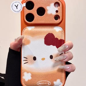 May include: Orange phone case with a Hello Kitty design. The case has a leather-like texture with a cartoon image of Hello Kitty, a red bow, and the words "Hello Kitty". The case has a camera cutout and a Y2CASE sticker.