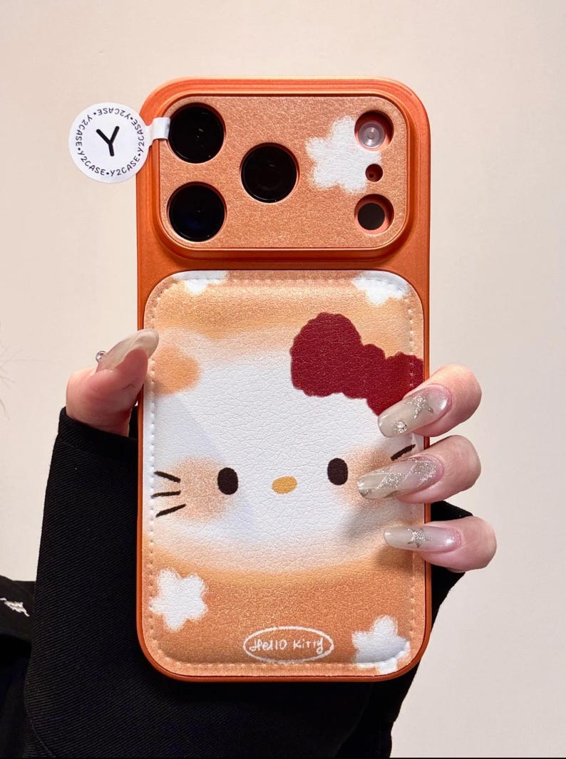 May include: Orange phone case with a Hello Kitty design. The case has a leather-like texture with a cartoon image of Hello Kitty, a red bow, and the words "Hello Kitty". The case has a camera cutout and a Y2CASE sticker.
