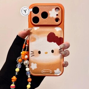 May include: Orange phone case with a Hello Kitty design, red bow, and cloud details. The case has a matching orange camera bump and a beaded charm strap. The words "Hello Kitty" are printed on the case.