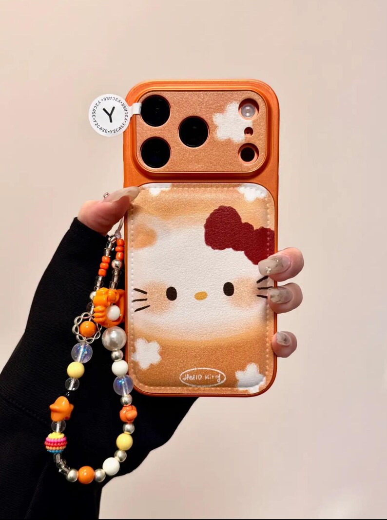 May include: Orange phone case with a Hello Kitty design, red bow, and cloud details. The case has a matching orange camera bump and a beaded charm strap. The words "Hello Kitty" are printed on the case.