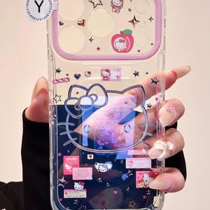 Pode incluir: Capa de telefone transparente com design Hello Kitty. A capa tem uma borda rosa e é decorada com estrelas, corações e gráficos Hello Kitty. O design inclui uma maçã com Hello Kitty.