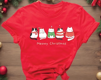 May include: A red t-shirt featuring a festive design with five cartoon cats dressed in holiday attire. The cats are wearing scarves, sweaters, and Santa hats. The text "Meowy Christmas" is printed below the cats.
