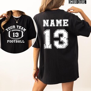 May include: Black t-shirt with white text and graphics. The front features a football graphic with the words "YOUR TEAM" above and "13" inside the football, with "FOOTBALL" below. The back displays "NAME" above and the number "13".