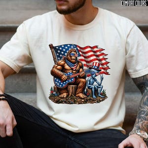 May include: A cream-colored t-shirt featuring a graphic of a brown Bigfoot playing a guitar and a blue alien sitting on tree stumps. Both figures wear American flag-themed sunglasses and accessories. A tattered American flag is in the background.