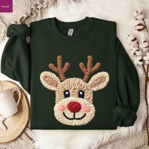 May include: A dark forest green crewneck sweatshirt featuring a textured, raised punch needle design of a reindeer face. The reindeer has light beige fur, brown antlers, dark eyes, a bright red nose, and pink cheeks on a soft fabric base.