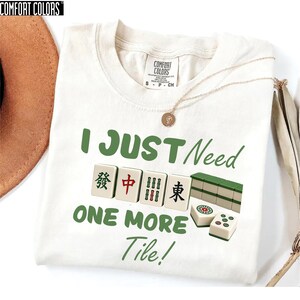 May include: Cream-colored t-shirt with the text "I JUST Need ONE MORE Tile!" in green. The design includes Mahjong tiles. A gold necklace with a pendant is draped across the shirt.