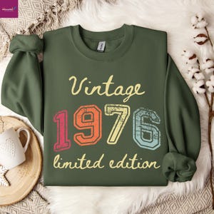 May include: Green sweatshirt with the text "Vintage 1976 limited edition" in a retro font. The numbers are in red, orange, yellow, and blue. The sweatshirt is folded on a white surface.