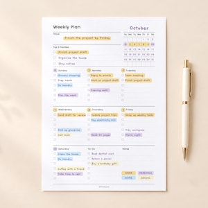 May include: A white weekly planner sheet with purple and yellow accents, featuring an October calendar, a focus section, top three priorities, and daily task lists for Sunday through Saturday, accompanied by a sleek cream-colored ballpoint pen.