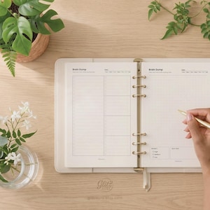 Pode incluir: Uma agenda de argolas aberta com p&aacute;ginas cor de creme com cabe&ccedil;alhos Brain Dump, um layout de grade de pontos e caixas de sele&ccedil;&atilde;o de prioridade. A agenda tem uma capa clara com argolas douradas e uma caneta dourada na p&aacute;gina direita.