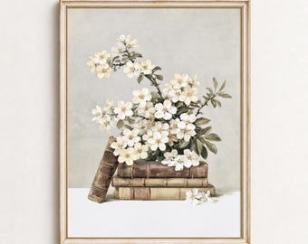 May include: A framed wall art print featuring a stack of three antique leather-bound books with a cluster of white apple blossoms resting on top. The composition is set against a soft, neutral gray background with a light-colored surface below.