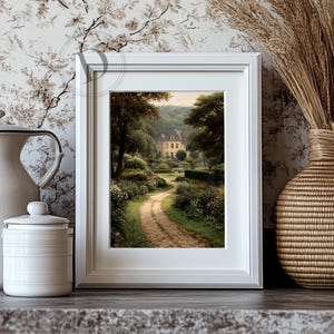 May include: A framed landscape print depicts a winding dirt path leading to a large house surrounded by lush greenery. The artwork is in a white frame, and the scene is filled with trees and flowers. The print is displayed on a gray surface.