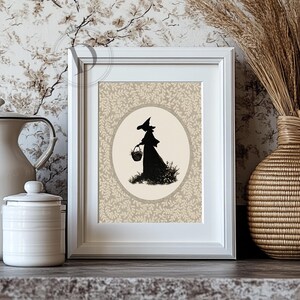 May include: Framed art print featuring a black silhouette of a witch carrying a basket, set against a beige oval and a patterned background. The artwork is displayed in a white frame, evoking a vintage or Halloween theme.