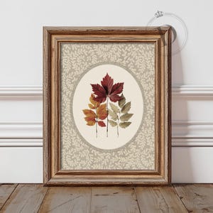 May include: Framed artwork featuring a central oval with colorful autumn leaves. The leaves range in color from deep red to yellow and green. The background has a light gray floral pattern, and the frame is a warm brown.