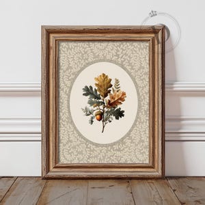 May include: Framed botanical print featuring an oak branch with green, gold, and brown leaves and an acorn. The artwork is set against a beige background with a repeating leaf pattern, enclosed in a wooden frame.