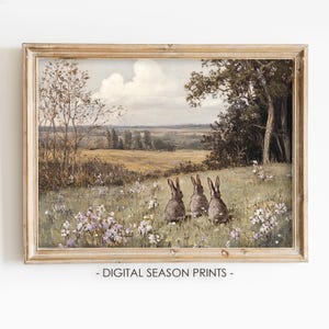 Könnte beinhalten: Ein Vintage-Gemälde mit drei braunen Kaninchen, die auf einer Wildblumenwiese sitzen. Die Kaninchen blicken auf eine ferne Landschaft mit blauem Himmel und flauschigen weißen Wolken. Das Gemälde ist in einem hellen Holzrahmen gerahmt.