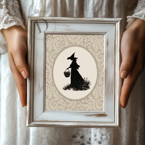 May include: A framed print featuring a black silhouette of a witch carrying a basket, set against a beige oval and a patterned background. The frame is white with a distressed finish, adding a vintage aesthetic. The artwork evokes a whimsical, Halloween theme.