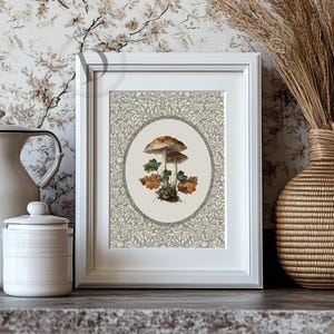 May include: A framed botanical print featuring two mushrooms with brown caps and green and orange leaves. The artwork is set against a gray and white floral patterned background, enclosed in a white frame. The print is displayed on a dark gray surface.