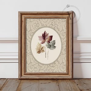 May include: A framed print depicting three stylized leaves in burgundy, green, and tan. The leaves are set against a cream oval, surrounded by a beige floral pattern. The frame is a warm, light brown.