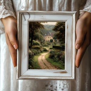 May include: A framed landscape print featuring a winding path leading to a large house. The print is in a white distressed wooden frame. The scene is filled with lush greenery and trees, creating a serene and picturesque view.