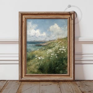 May include: A framed landscape painting depicts a coastal scene with a blue sky and fluffy white clouds. The foreground features green grass and white wildflowers. The artwork is in a wooden frame.
