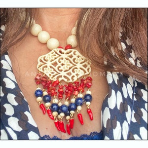 May include: A white carved pendant necklace with red and blue beads. The pendant is a large, intricate design with a filigree pattern. The beads are strung on a gold chain.