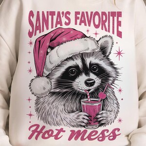 May include: White sweatshirt with a graphic of a raccoon wearing a pink Santa hat and holding a glass of pink liquid with a straw. The text "Santa's Favorite Hot Mess" is printed on the sweatshirt.