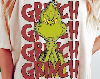 May include: Cream-colored t-shirt featuring a green Grinch character with the word "GRINCH" in red, stacked three times. The Grinch has a grumpy expression. The shirt is a casual, oversized fit.