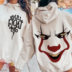 May include: Cream-colored hoodie with a front graphic that reads "YOU'LL FLOAT TOO" in a stylized font. The back features a red and black clown face design. The hoodie has a drawstring hood and a front pocket.