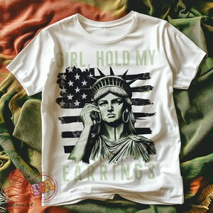 May include: White t-shirt featuring a graphic of the Statue of Liberty holding a phone and the text "Girl, Hold My Earrings".
