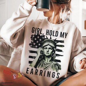 May include: A white sweatshirt featuring a graphic of the Statue of Liberty holding a phone to her ear. The graphic is set against an American flag with the text "Girl, Hold My Earrings" printed below.