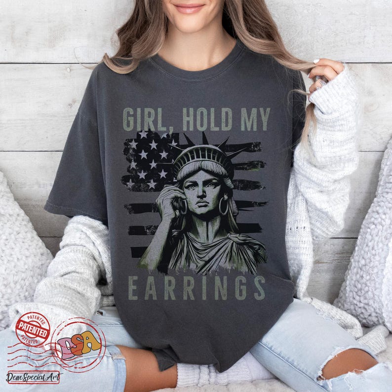 May include: A dark grey t-shirt featuring a graphic of the Statue of Liberty holding a large earring. The graphic is superimposed on an American flag. The text "Girl, Hold My Earrings" is printed on the shirt.