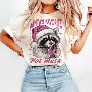 May include: A white t-shirt with a pink graphic of a raccoon wearing a Santa hat and drinking a red drink with a straw. The text "Santa's Favorite Hot Mess" is printed in pink on the shirt.