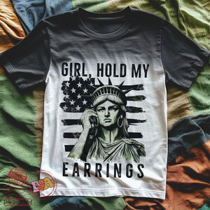 May include: A black and white t-shirt featuring a graphic of the Statue of Liberty holding a phone and the text "Girl, hold my earrings" on a red, white and blue American flag background.
