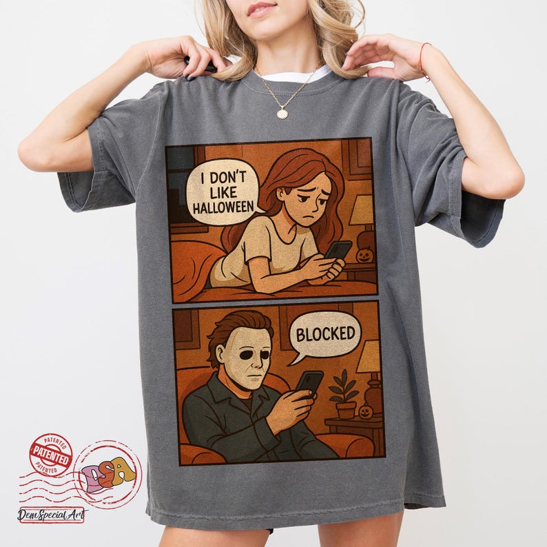 K&ouml;nnte beinhalten: Dunkelgraues T-Shirt mit einem zweiteiligen Comic. Das obere Panel zeigt eine Frau, die textet, mit dem Text "I DON'T LIKE HALLOWEEN". Das untere Panel zeigt einen Mann mit Maske, der textet, mit dem Text "BLOCKED".