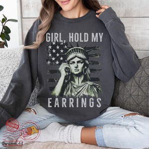 May include: A dark grey sweatshirt featuring a graphic of the Statue of Liberty holding a large earring. The graphic is superimposed on an American flag background. The text "Girl, Hold My Earrings" is printed on the sweatshirt.