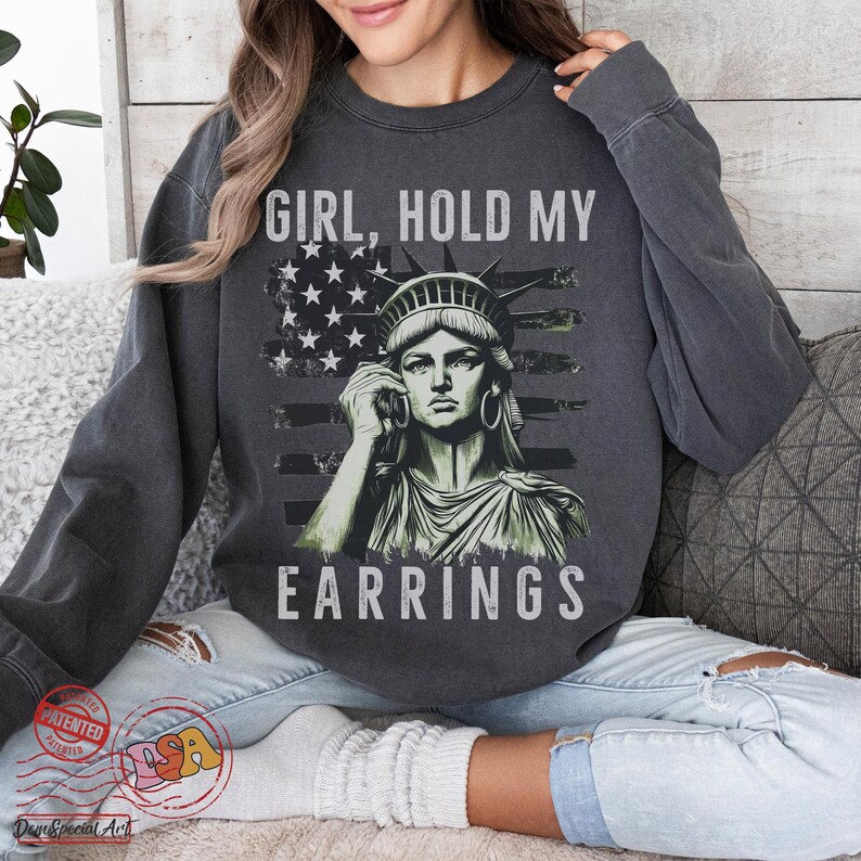 May include: A dark grey sweatshirt featuring a graphic of the Statue of Liberty holding a large earring. The graphic is superimposed on an American flag background. The text "Girl, Hold My Earrings" is printed on the sweatshirt.