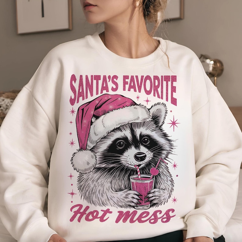 May include: White sweatshirt with a graphic of a raccoon wearing a pink Santa hat and holding a glass of pink liquid with a straw. The text "Santa's Favorite Hot Mess" is printed on the sweatshirt.