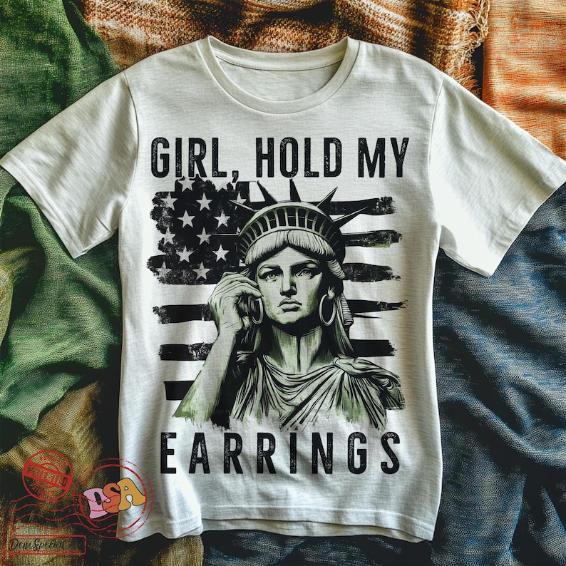May include: White t-shirt featuring a graphic of the Statue of Liberty holding a phone, with the text "Girl, Hold My Earrings" printed on it. The Statue of Liberty is in front of an American flag.