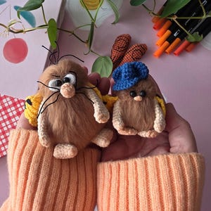 May include: Two handmade, plush, brown creatures with crocheted details. One has a blue cap, the other has long whiskers. Both have white crocheted arms and legs, and yellow wings. The creatures are held in a person's hands, wearing an orange jumper.