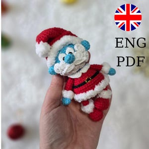 May include: A crocheted Smurf dressed as Santa Claus, featuring a red and white hat and suit with a black belt. The Smurf has blue skin and a white beard. The image includes the text "ENG PDF" and a British flag.