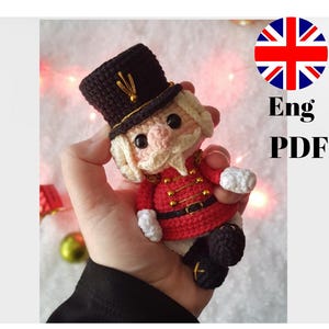 Puede incluir: Un muñeco cascanueces de crochet con un sombrero negro, una chaqueta roja y guantes blancos. El muñeco sostiene un pequeño objeto blanco en su mano derecha.