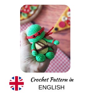 Puede incluir: Una figura de ganchillo verde hecha a mano de una Tortuga Ninja, con una cinta roja en la cabeza, ojos negros y un caparazón marrón. La figura se sostiene en una mano. El texto "Crochet Pattern in ENGLISH" está debajo.