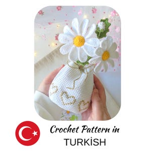 May include: A white crocheted vase with gold heart accents holds crocheted daisies with white petals and yellow centers. The vase is tied with a gold ribbon. The text "Crochet Pattern in TURKISH" is below the vase.