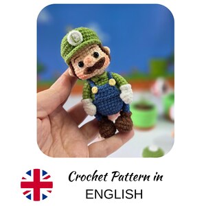 Può includere: Bambola di Luigi all'uncinetto, con cappello verde, baffi marroni e tuta blu. La bambola è tenuta in mano, evidenziando il dettagliato lavoro all'uncinetto. Lo sfondo è una scena di videogioco sfocata. Circa 10 cm di altezza.