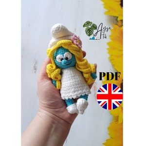 Könnte beinhalten: Gehäkelte Schlumpfette-Puppe mit einem weißen Kleid, gelben Haaren und einer rosa Blume. Die Puppe wird in einer Hand gehalten. Das Bild enthält auch den Text "Ayse Tik" und "PDF".